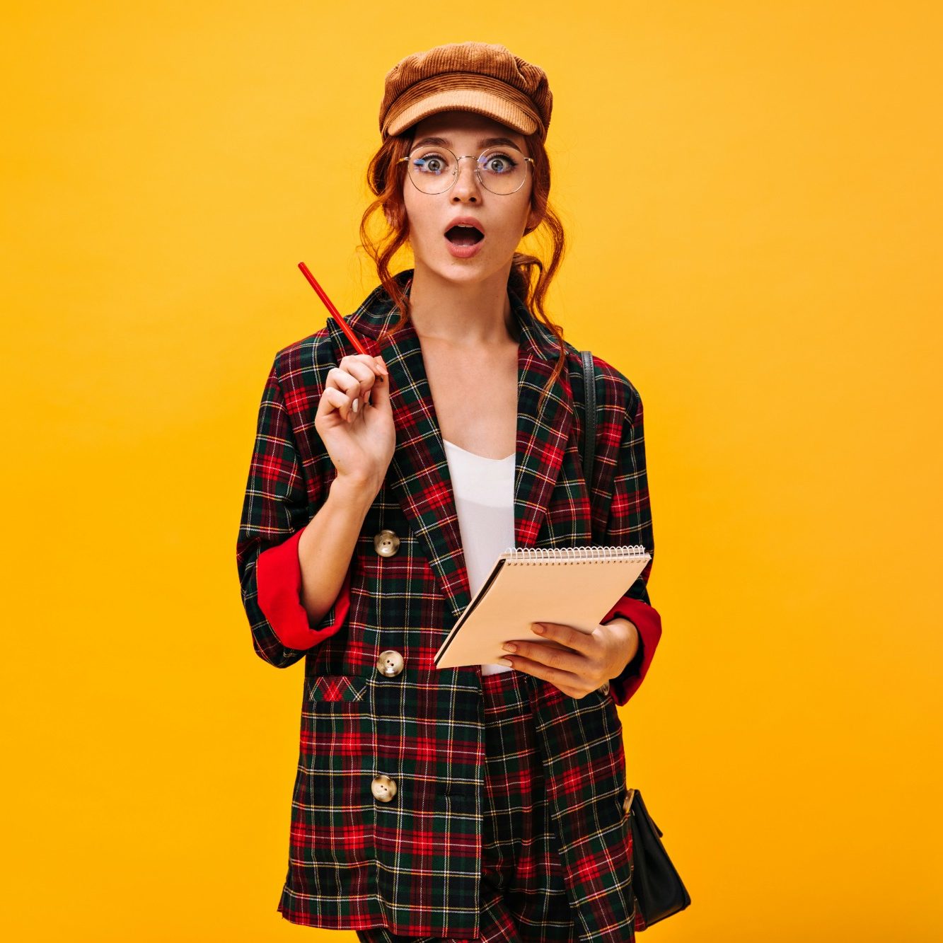 Femme surprise avec un stylo et un carnet, vêtue d'un blazer à carreaux sur fond jaune.