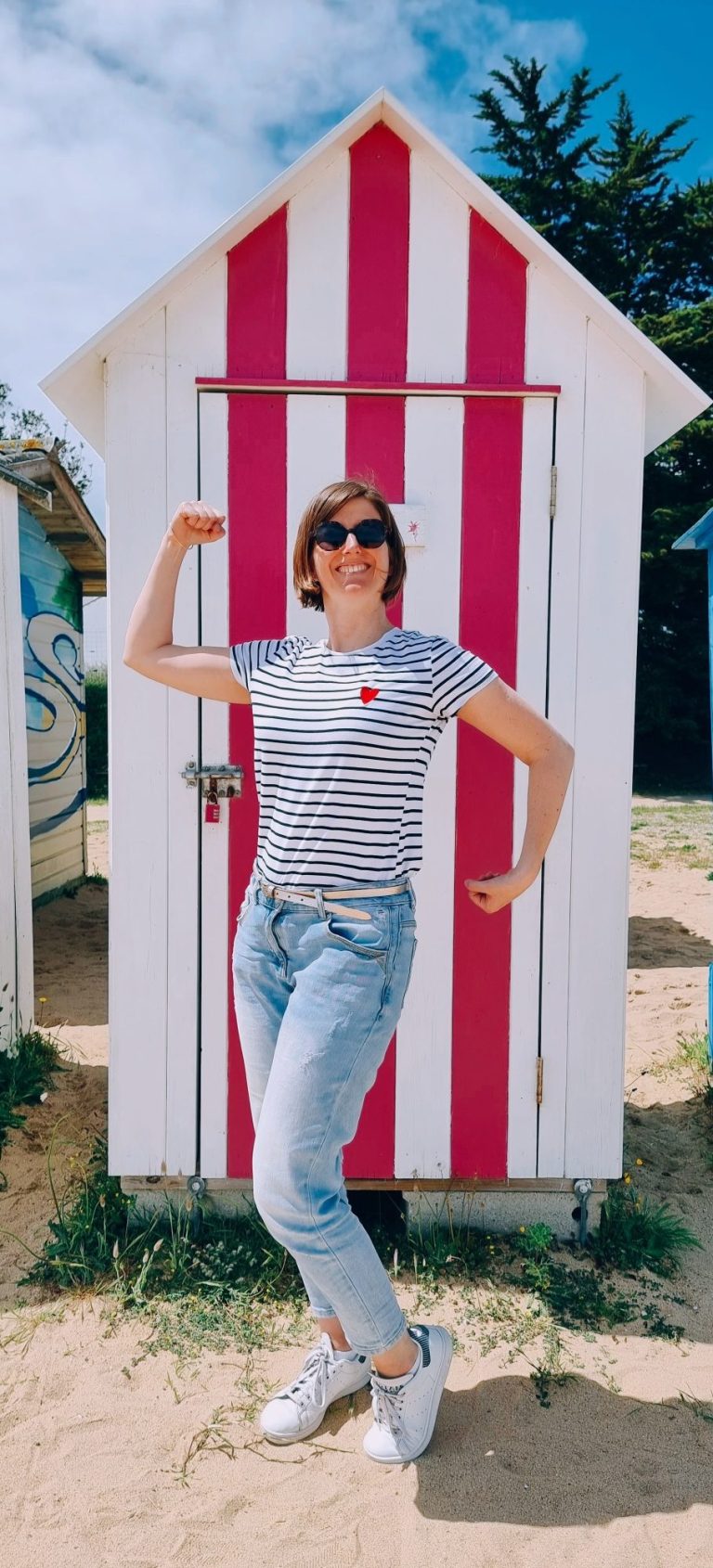 Femme en t-shirt rayé et jeans devant des cabines de plage colorées.
